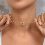 A close-up lifestyle shot of a model wearing a delicate gold chain necklace with two small charms, holding the chain out with both hands to show its scale.