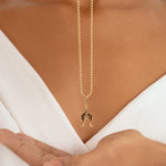 A close-up of a model wearing a delicate gold chain necklace with a small house-shaped pendant, styled with a white V-neck top.