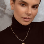 A close-up of a woman wearing a delicate gold chain necklace with small white pearls and a small gold pendant, paired with a black turtleneck sweater.