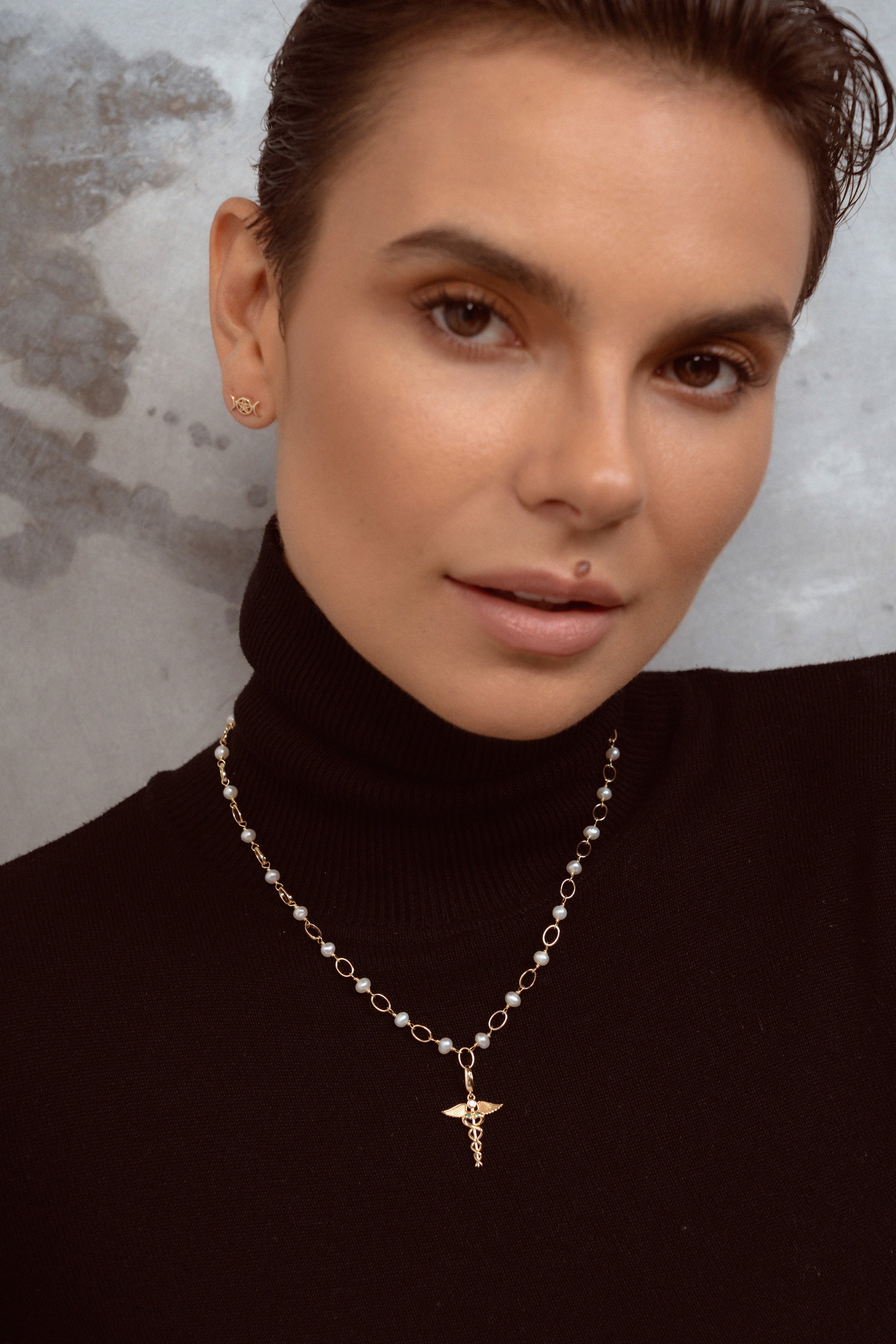 A close-up of a woman wearing a delicate gold chain necklace with small white pearls and a small gold pendant, paired with a black turtleneck sweater.