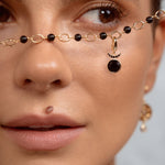 Close-up of a model wearing a delicate gold chain necklace with black beads and a crescent moon pendant draped across her nose, with a matching earring visible on her ear.