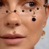Close-up of a model wearing a delicate gold chain necklace with black beads and a crescent moon pendant draped across her nose, with a matching earring visible on her ear.