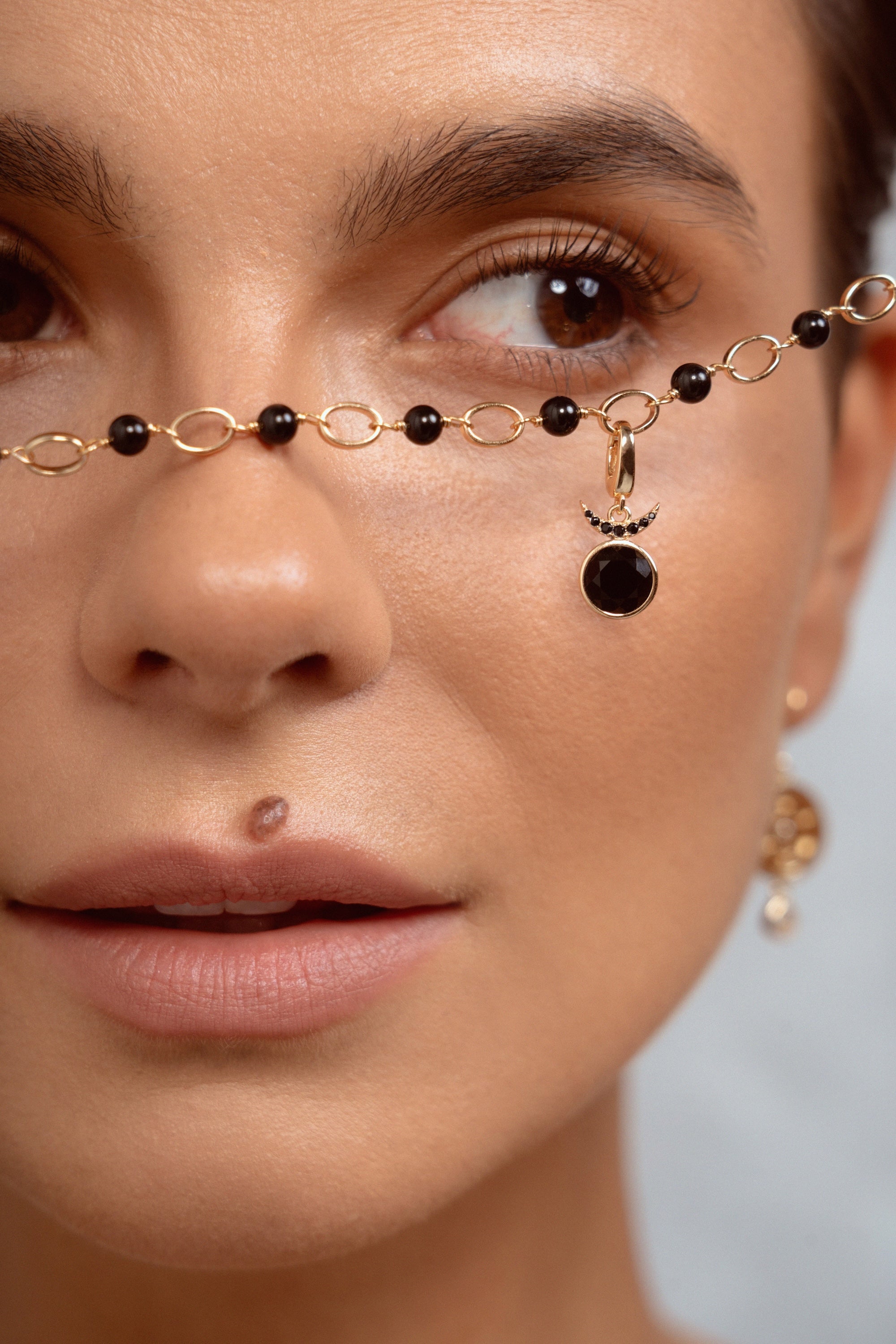 Close-up of a model wearing a delicate gold chain necklace with black beads and a crescent moon pendant draped across her nose, with a matching earring visible on her ear.