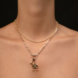 A close-up of a model's neck and upper chest wearing a layered gold necklace with a small house-shaped pendant, against a dark background.