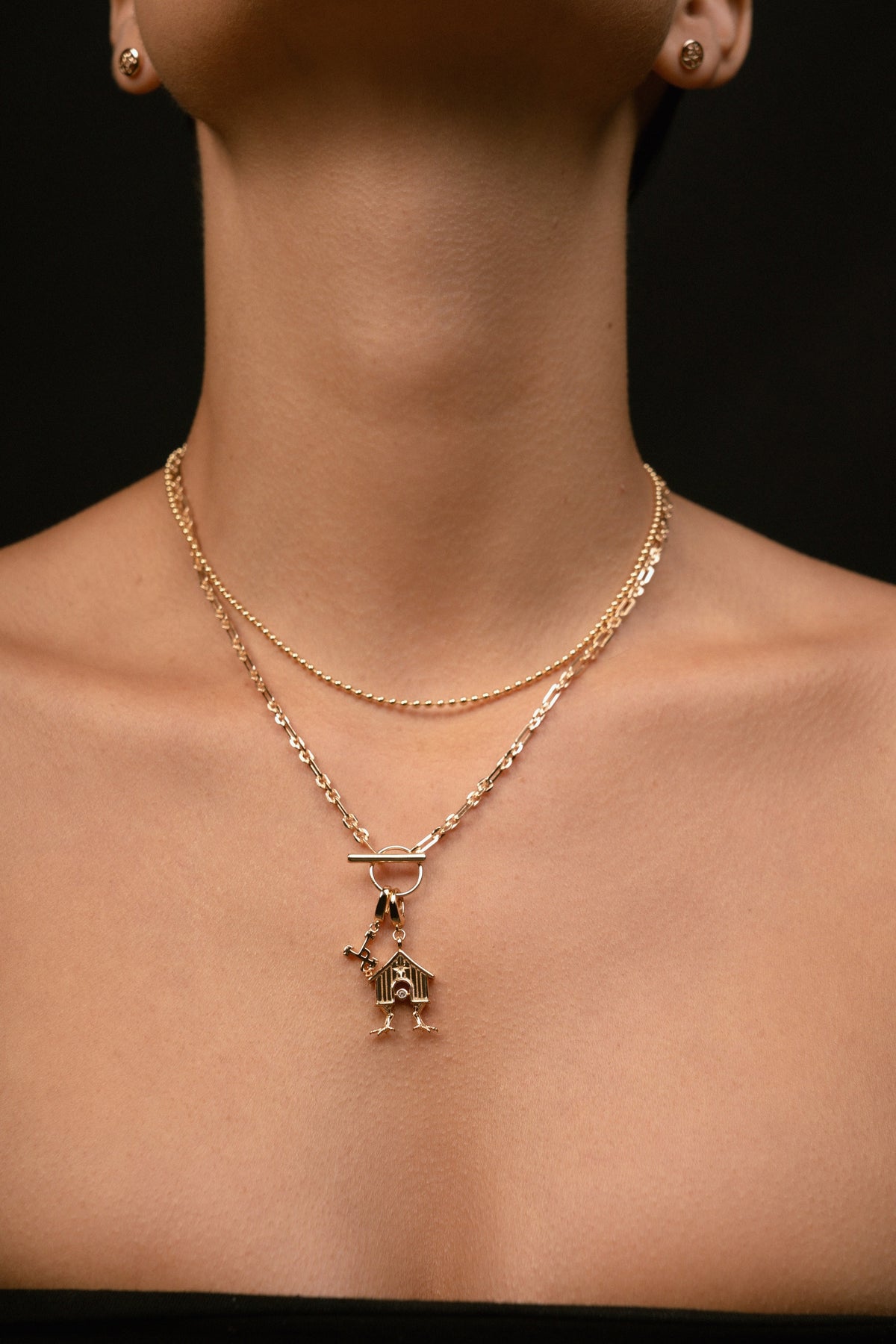 A close-up of a model's neck and upper chest wearing a layered gold necklace with a small house-shaped pendant, against a dark background.