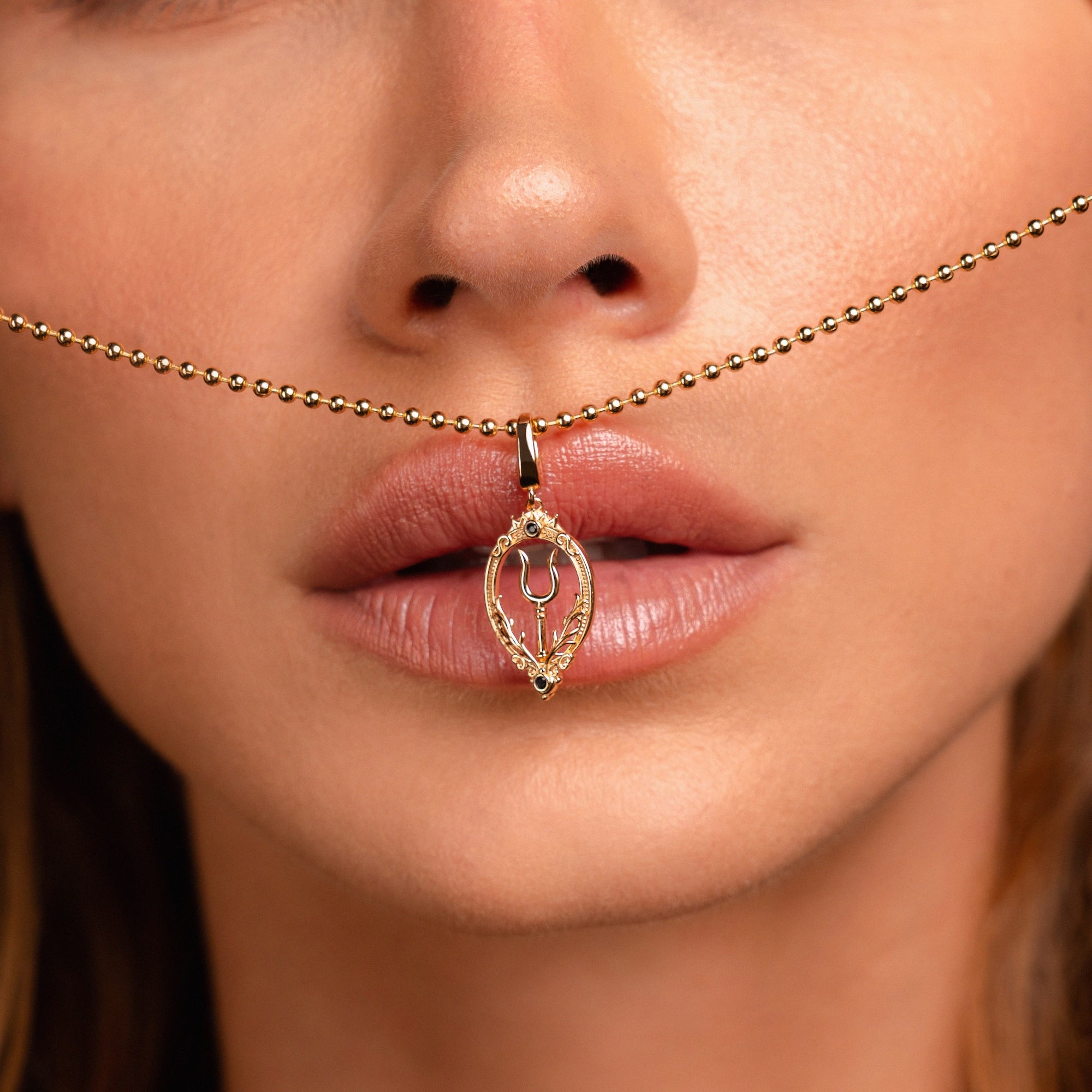 A close-up of a woman’s face with a gold necklace featuring a bident pendant held between her lips and nose.