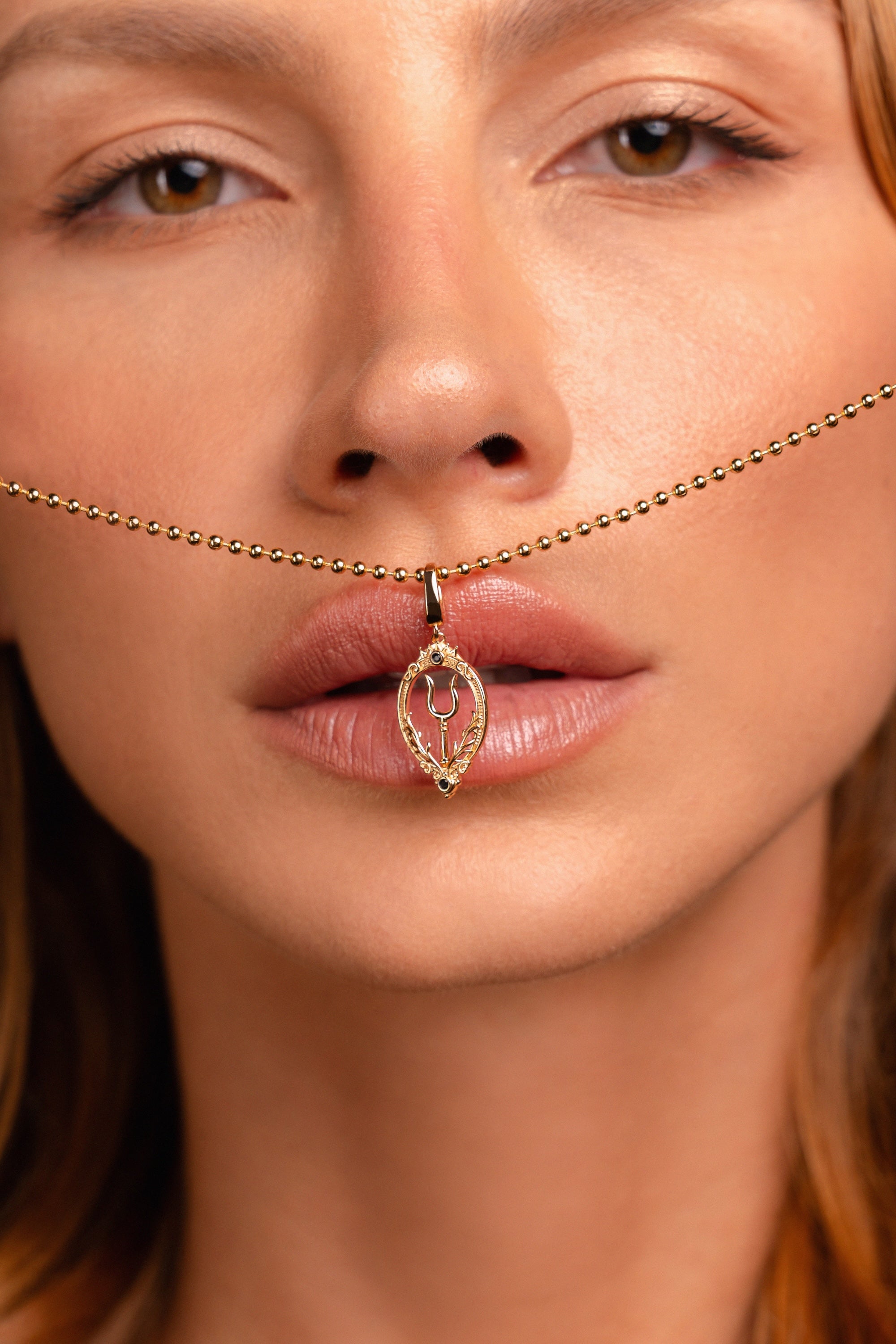 A close-up of a woman’s face with a gold necklace featuring a bident pendant held between her lips and nose.