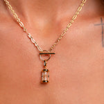 The Hourglass Charm, a gold chain necklace with an hourglass pendant, is worn around the neck. Part of a tattooed cross appears on the skin near the right collarbone.