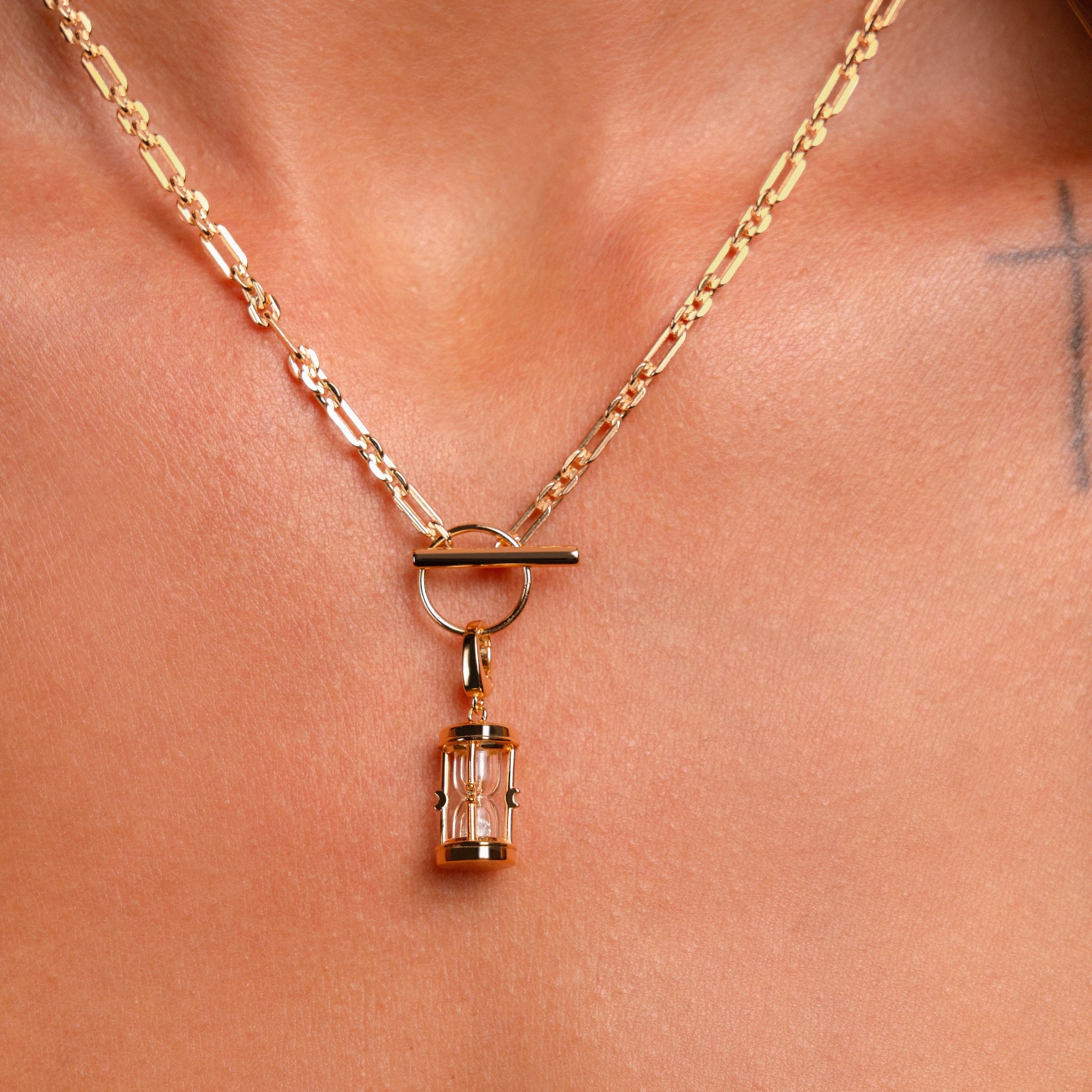 The Hourglass Charm, a gold chain necklace with an hourglass pendant, is worn around the neck. Part of a tattooed cross appears on the skin near the right collarbone.