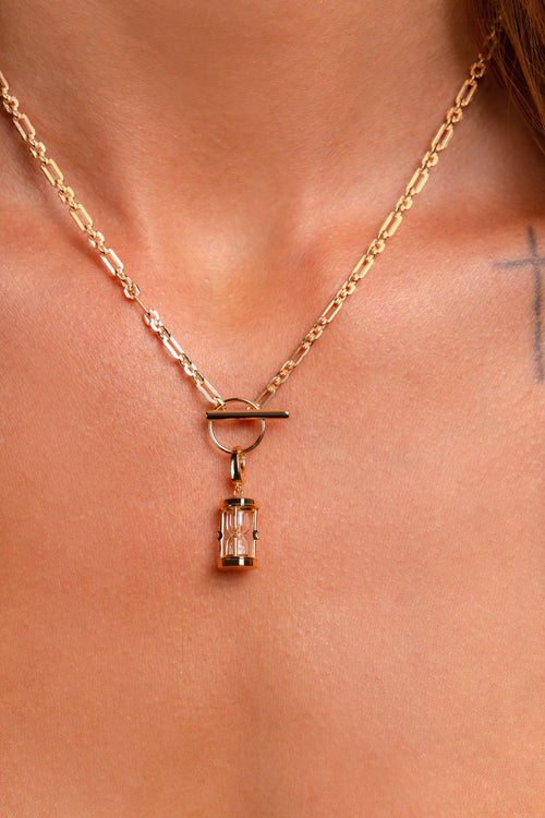 The Hourglass Charm, a gold chain necklace with an hourglass pendant, is worn around the neck. Part of a tattooed cross appears on the skin near the right collarbone.