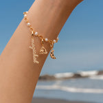 Gold charm bracelet with pearls featuring a script ‘magickal’ charm, a gold triquetra charm, and a gold crescent moon charm with black and white stones, photographed against a beach background