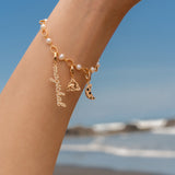 Gold charm bracelet with pearls featuring a script ‘magickal’ charm, a gold triquetra charm, and a gold crescent moon charm with black and white stones, photographed against a beach background