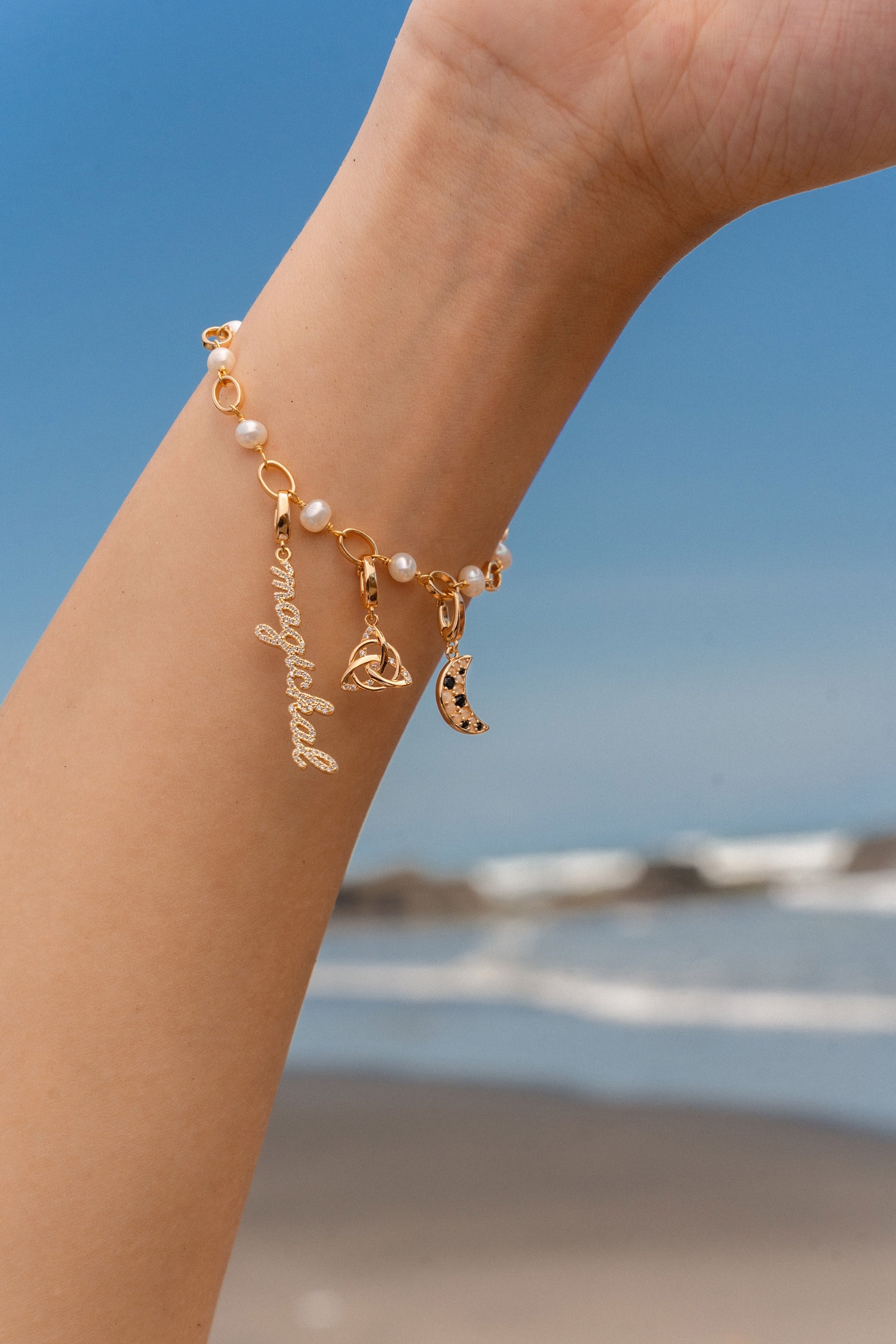 Gold charm bracelet with pearls featuring a script ‘magickal’ charm, a gold triquetra charm, and a gold crescent moon charm with black and white stones, photographed against a beach background