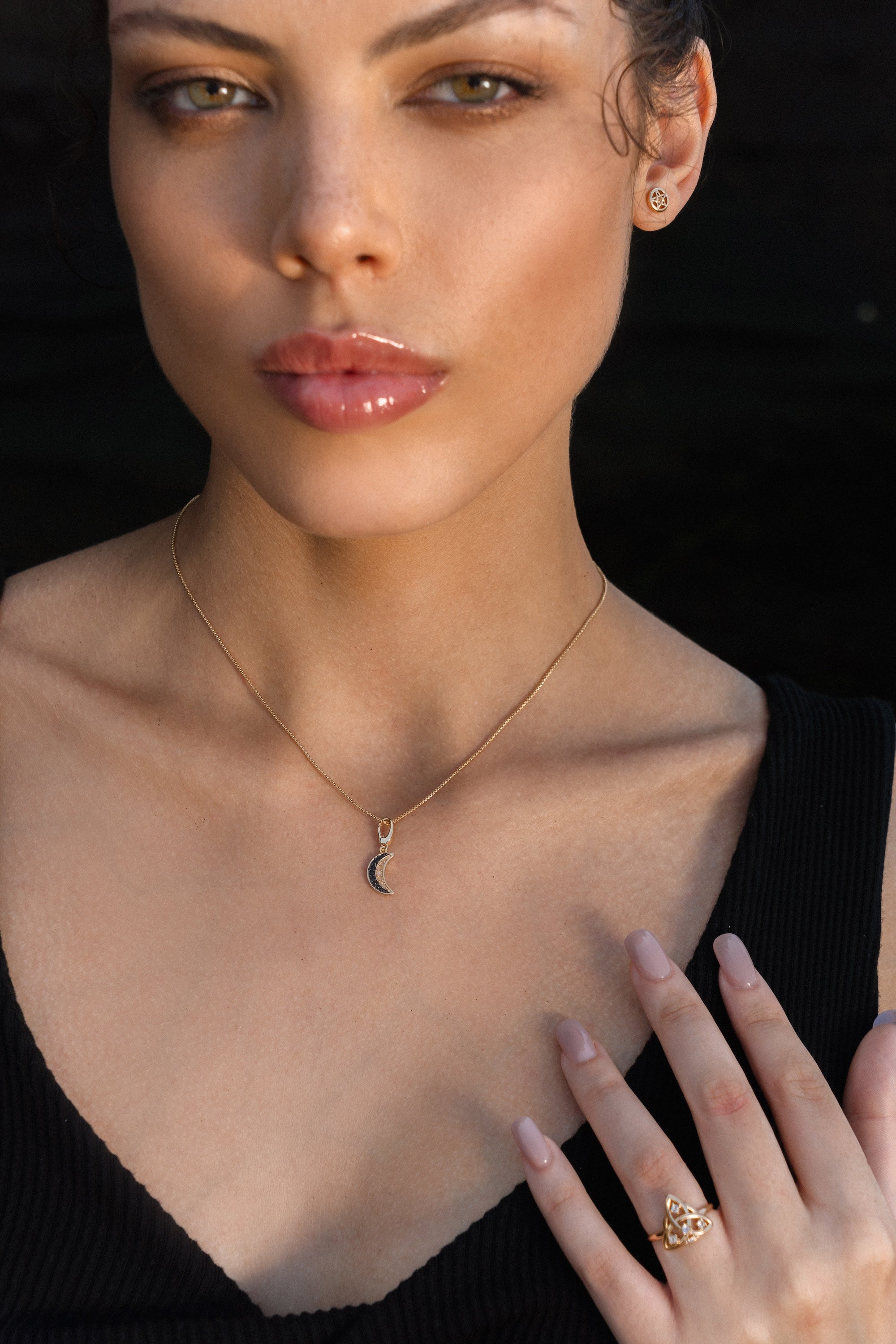 Woman wearing a gold crescent moon necklace, gold pentacle stud earrings, and a gold Triquetra ring with an intricate design.