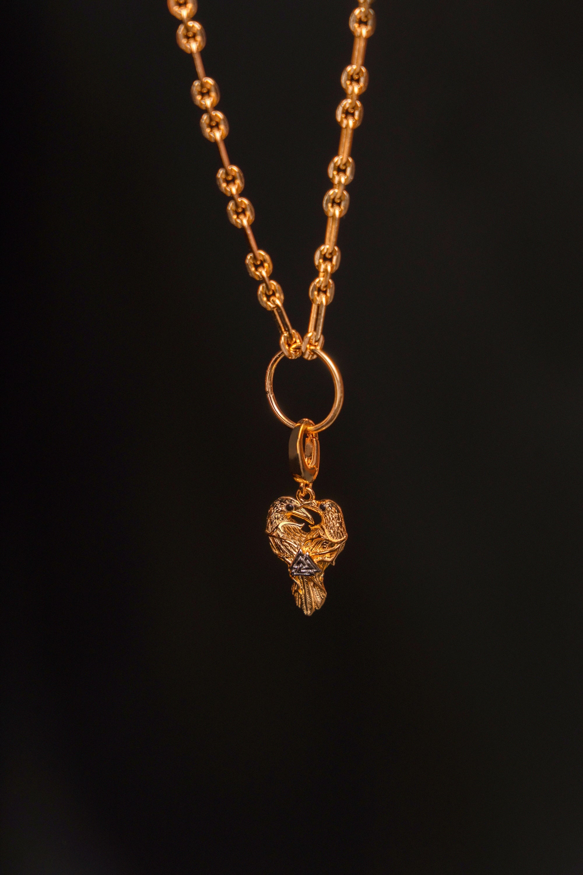 Preorder: Odin Charm (Ships end October 2026)—a gold chain necklace with a textured link and detailed heart pendant featuring two birds, inspired by Norse mythology, shown on a dark background.