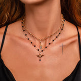 A close-up of a model wearing a layered necklace set. The top layer features a candle charm on a gold chain with small black beads, while the bottom layer is a gold chain with a gold bat pendant.