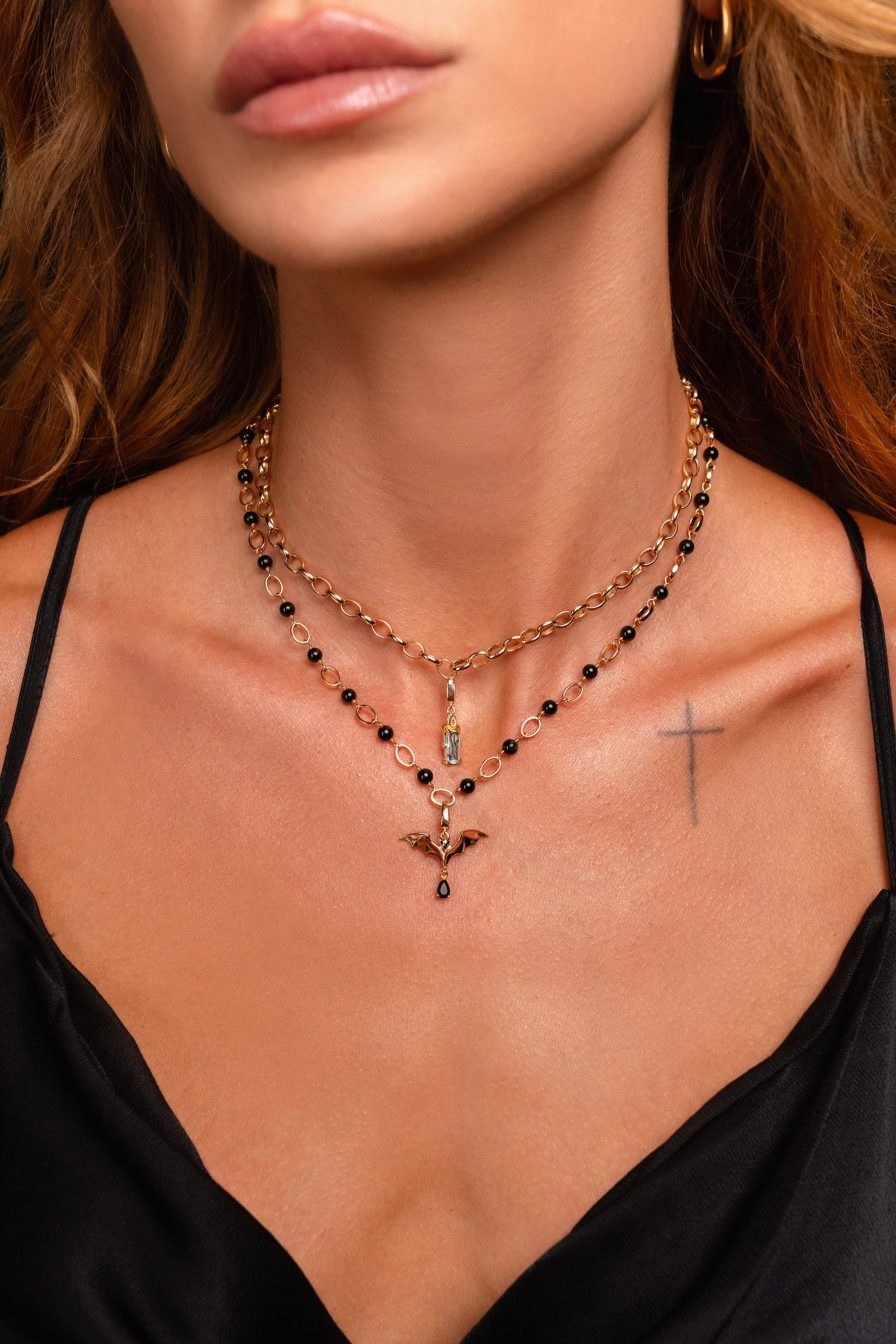 A close-up of a model wearing a layered necklace set. The top layer features a candle charm on a gold chain with small black beads, while the bottom layer is a gold chain with a gold bat pendant.
