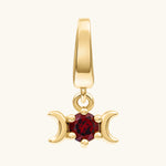 A gold pendant charm featuring a central round red gemstone flanked by two crescent moons, suspended from a polished gold bail, shown on a light cream background.