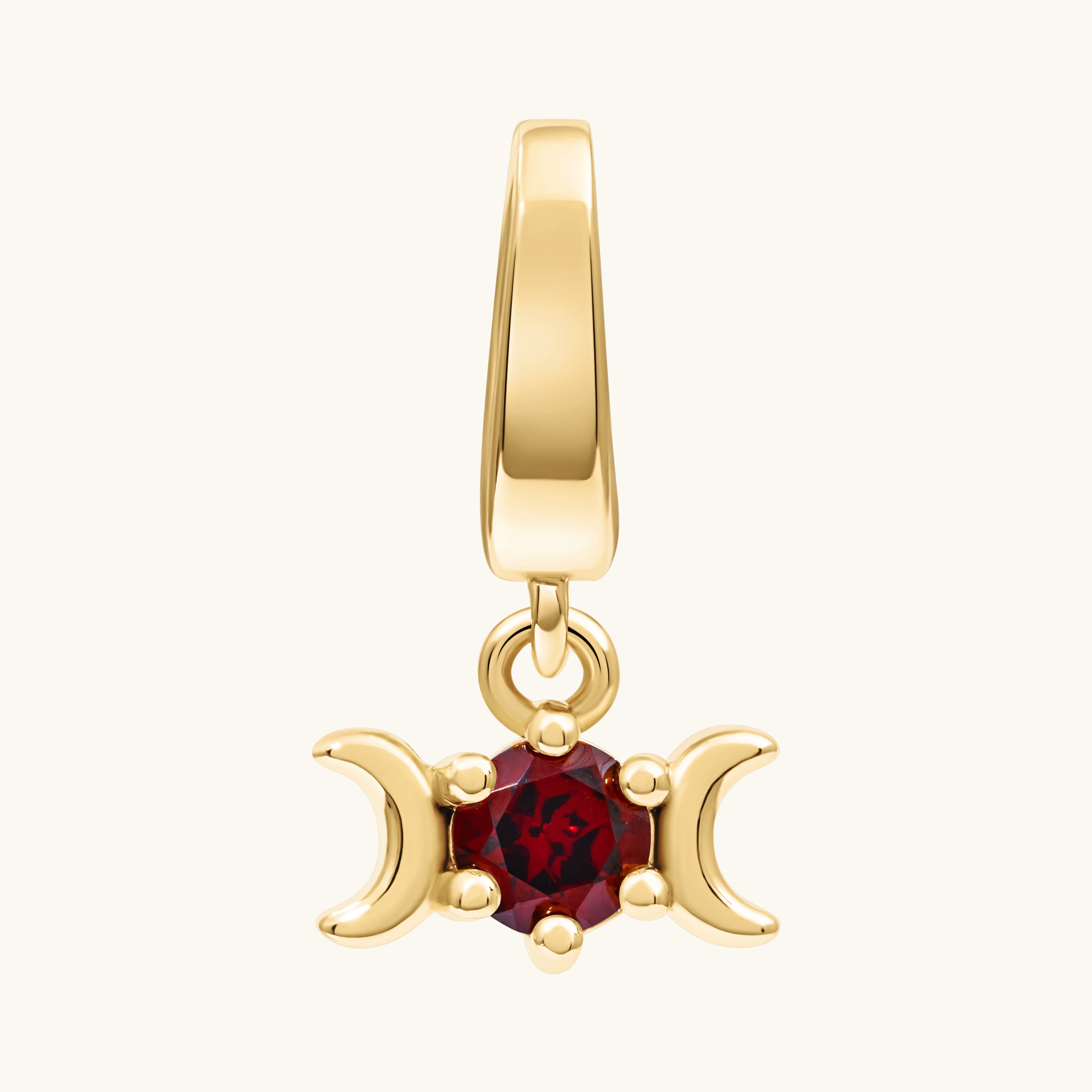 A gold pendant charm featuring a central round red gemstone flanked by two crescent moons, suspended from a polished gold bail, shown on a light cream background.