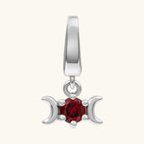 A silver triple moon pendant charm featuring a central faceted red gemstone, set against a plain off-white background.