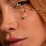 Close-up of a model's face holding up a gold chain necklace with a dangling Witch's Knot shaped charm studded with clear stones.
