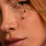 Close-up of a model's face holding up a gold chain necklace with a dangling Witch's Knot shaped charm studded with clear stones.