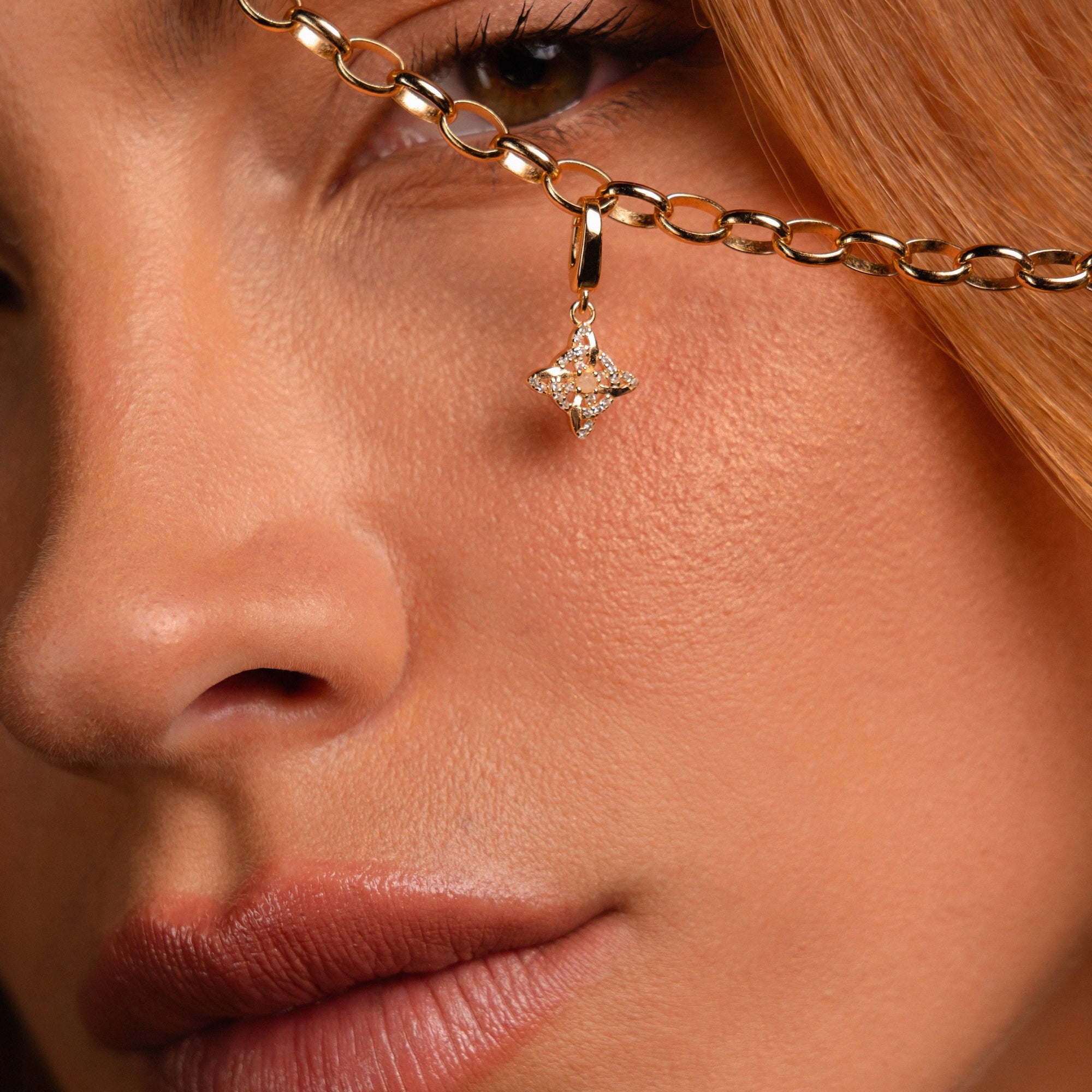 Close-up of a model's face holding up a gold chain necklace with a dangling Witch's Knot shaped charm studded with clear stones.