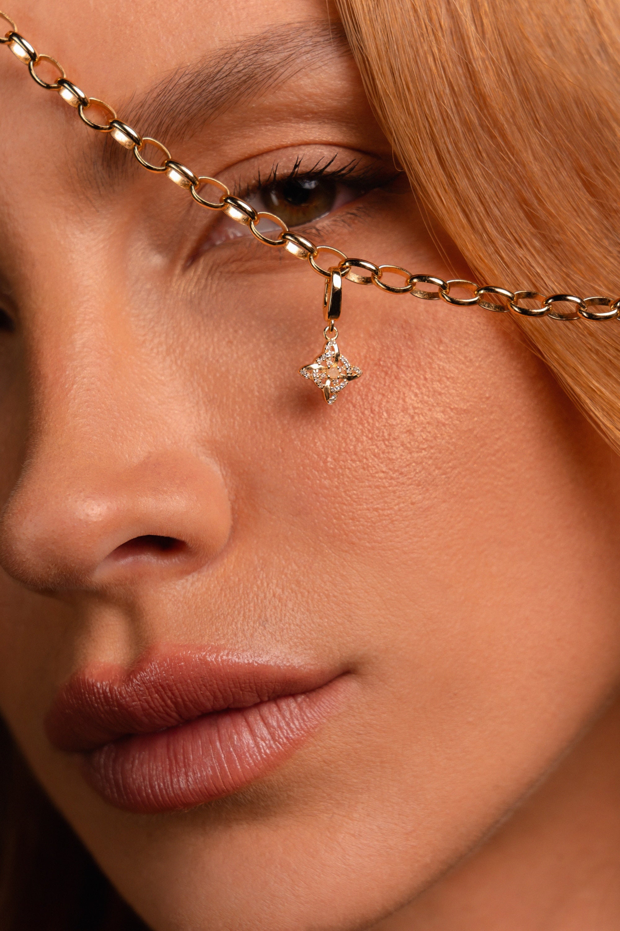 Close-up of a model's face holding up a gold chain necklace with a dangling Witch's Knot shaped charm studded with clear stones.
