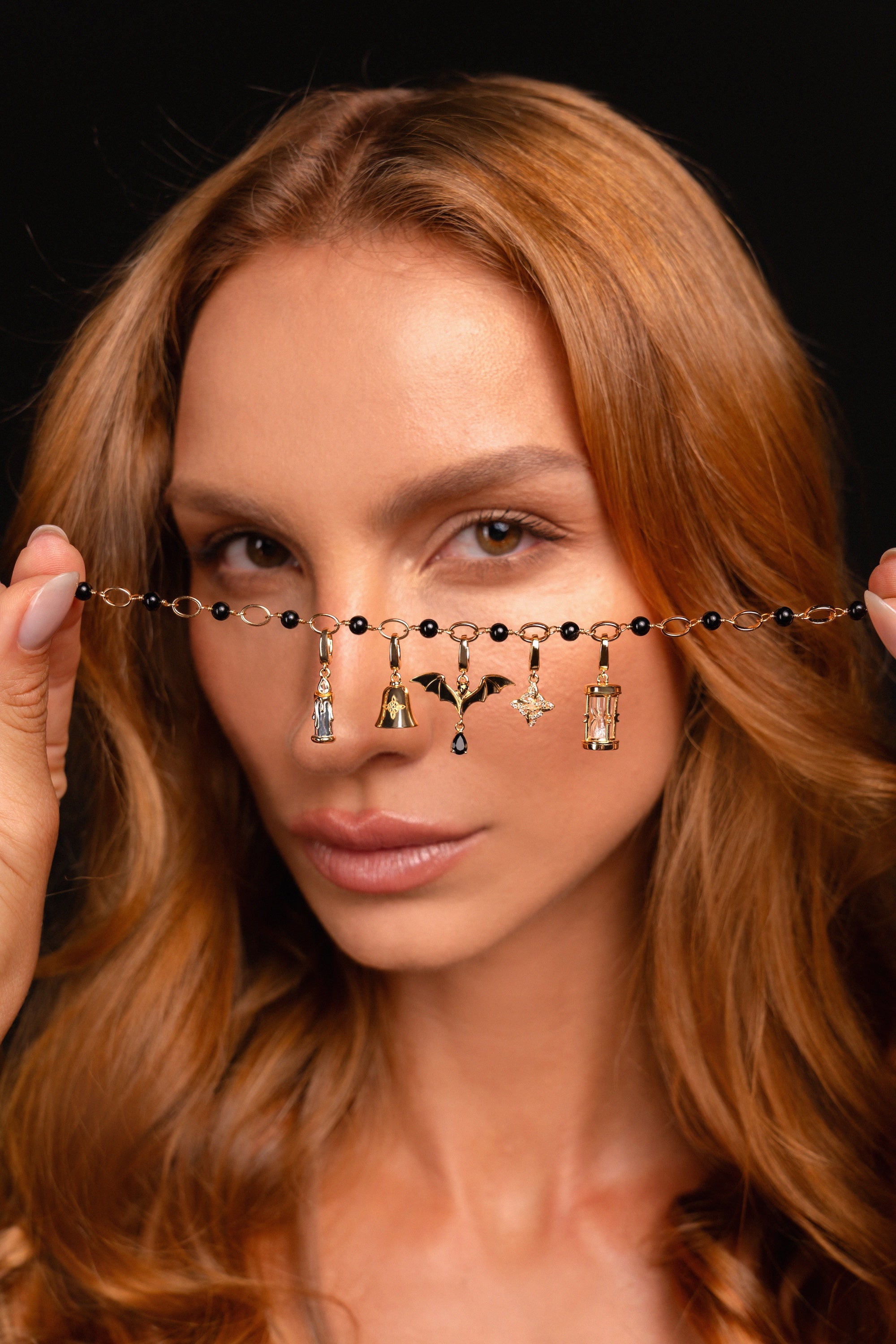 A close-up of a woman holding a delicate beaded chain across her eyes, featuring small black beads and dangling charms including a bat, a candle, and a small bell pendant, against a solid black background.