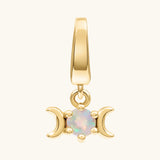A gold pendant charm featuring a triple moon symbol surrounding a round iridescent white opal gemstone, shown on a light cream background.