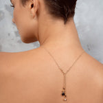 A woman shown from the back wearing a delicate gold Y-necklace with a small black and clear stone pendant, set against a textured gray background.