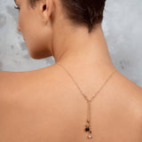 A woman shown from the back wearing a delicate gold Y-necklace with a small black and clear stone pendant, set against a textured gray background.