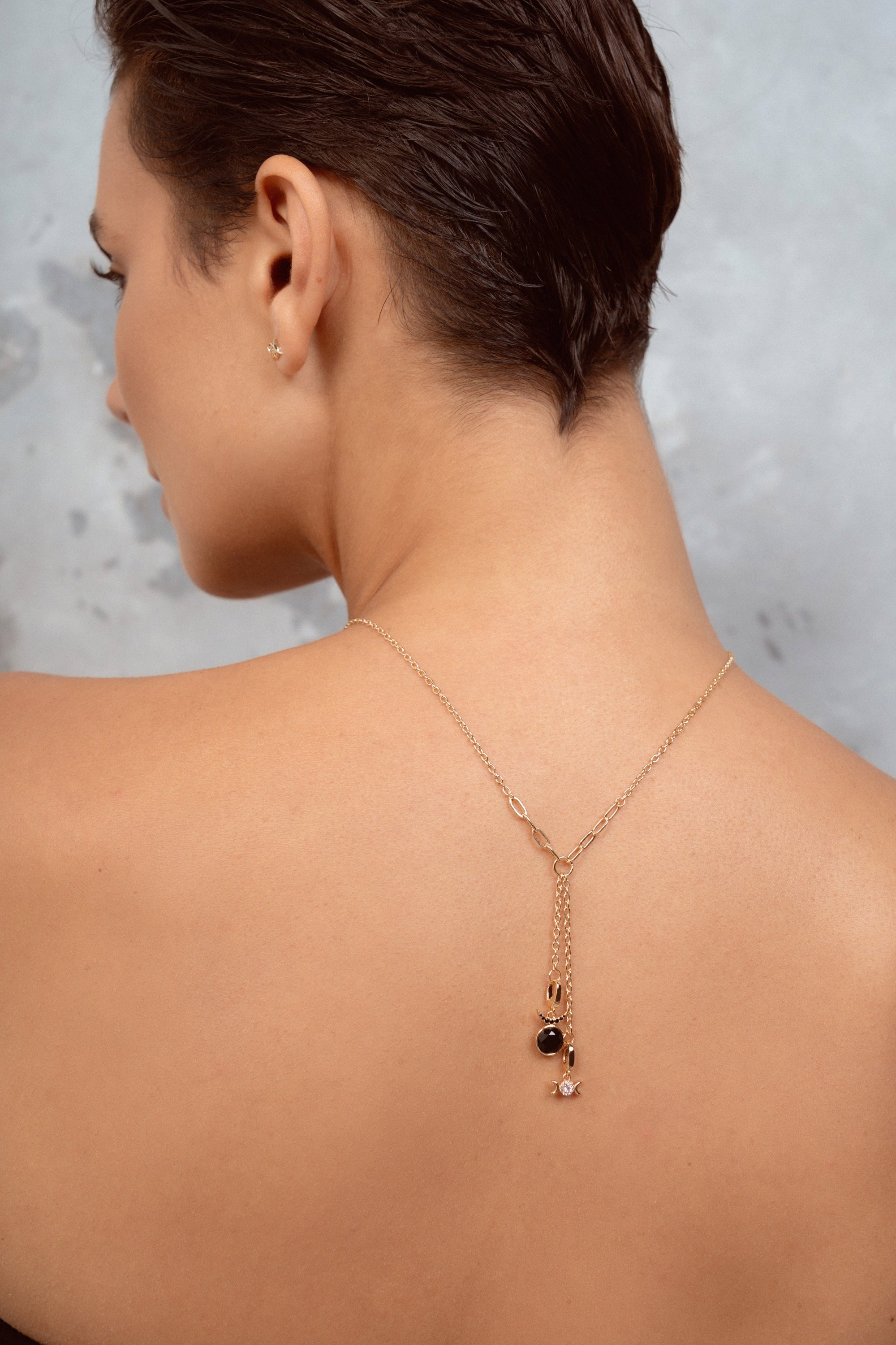 A woman shown from the back wearing a delicate gold Y-necklace with a small black and clear stone pendant, set against a textured gray background.