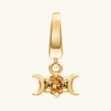 A gold pendant charm featuring a triple moon symbol surrounding a round iridescent yellow citrine gemstone, shown on a light cream background.