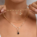 A close-up of a model wearing a layered gold necklace set, including a delicate chain choker with small dangling charms and a longer chain with a small black oval pendant, held gently by both hands.