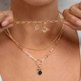 A close-up of a model wearing a layered gold necklace set, including a delicate chain choker with small dangling charms and a longer chain with a small black oval pendant, held gently by both hands.