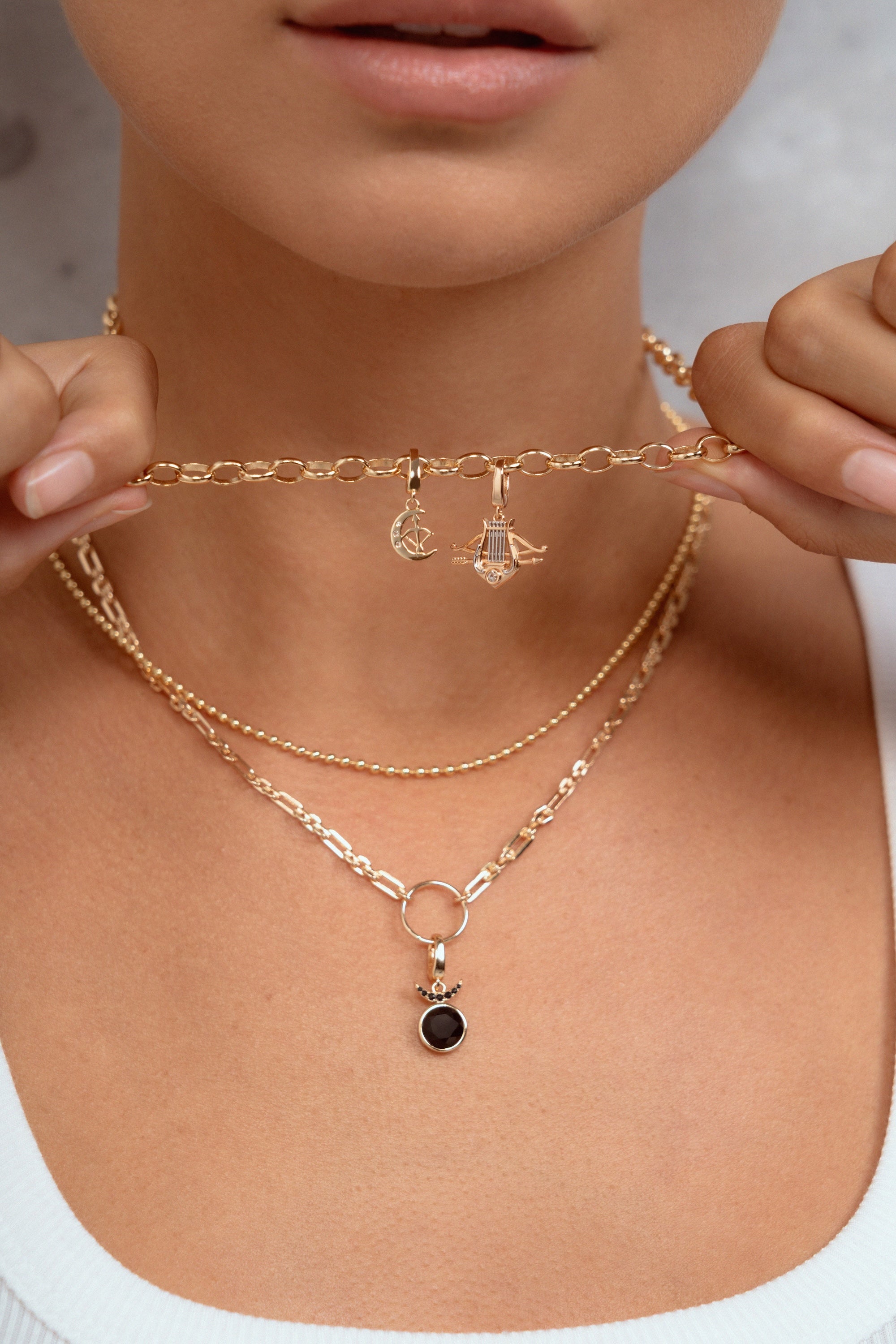 A close-up of a model wearing a layered gold necklace set, including a delicate chain choker with small dangling charms and a longer chain with a small black oval pendant, held gently by both hands.