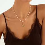 A close-up of a model wearing a delicate gold necklace with a small house-shaped pendant, styled with a black lace-trimmed camisole top against a clean white background.