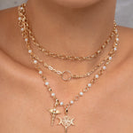 A close-up of a model wearing a layered gold necklace set with white pearl accents and two distinct pendants: a  key, and a caduceus symbol.