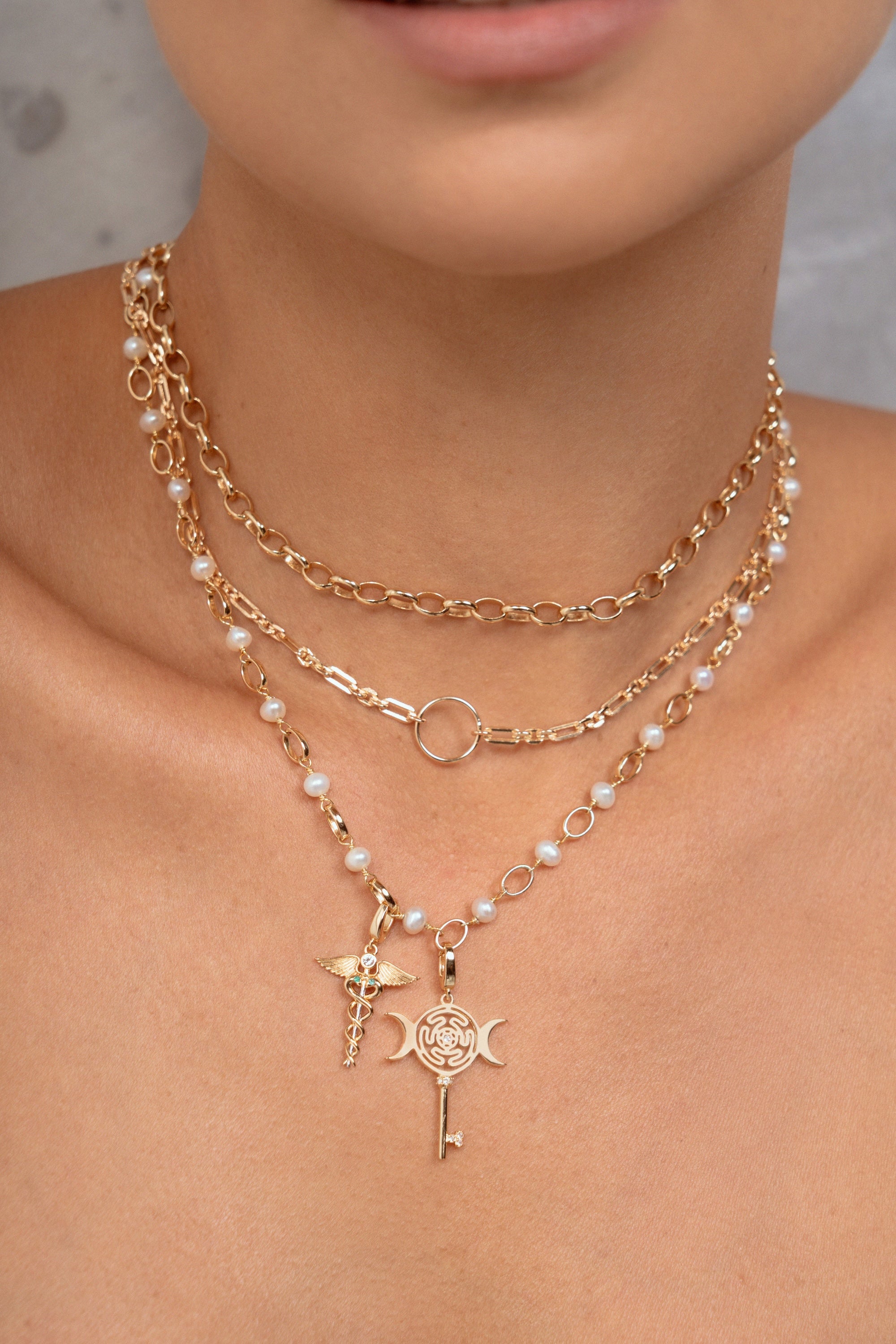 A close-up of a model wearing a layered gold necklace set with white pearl accents and two distinct pendants: a  key, and a caduceus symbol.