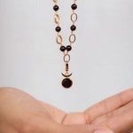 A delicate necklace with a gold chain featuring alternating dark black beads and oval links, hanging above an open hand against a light gray background.