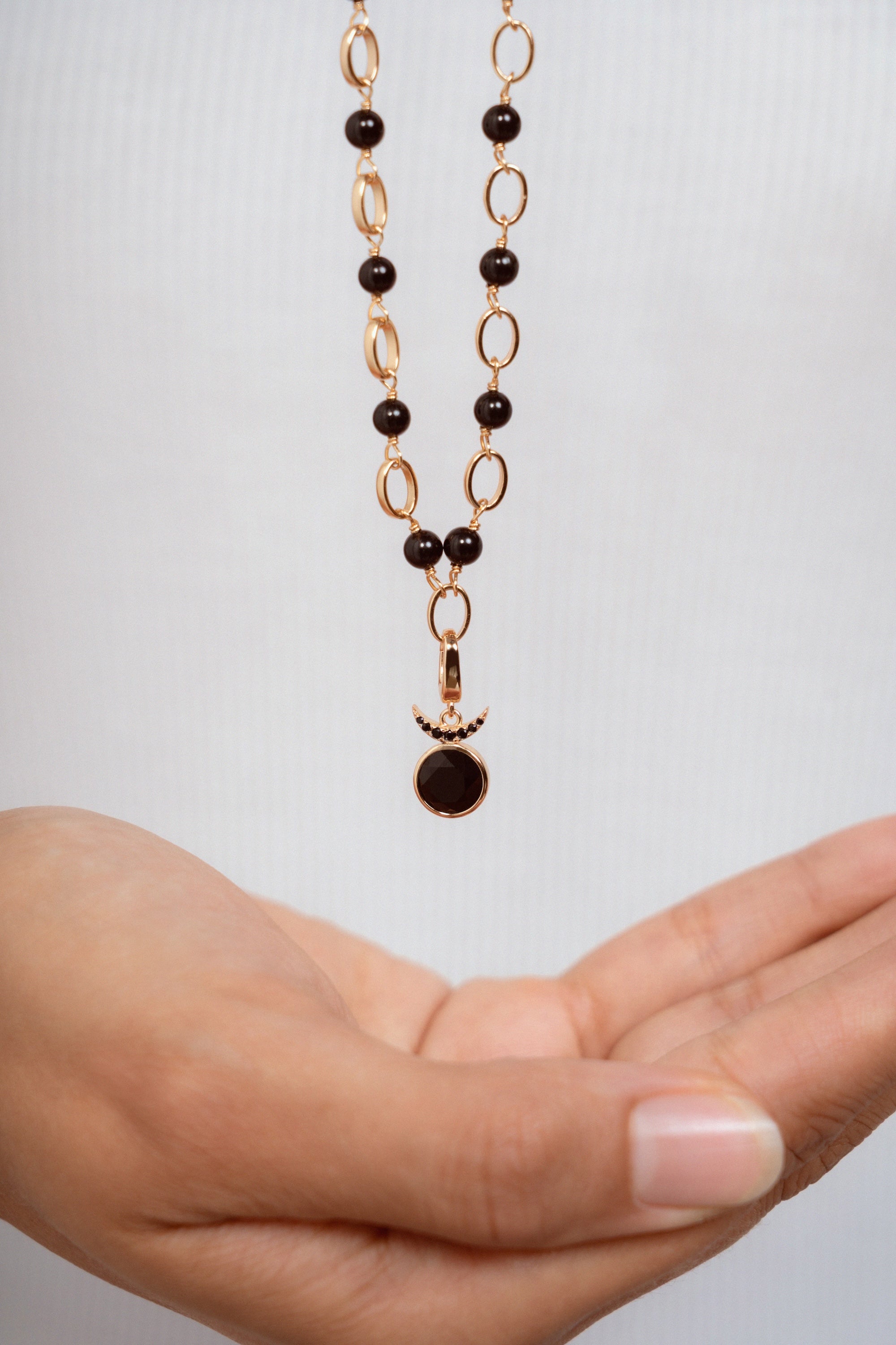 A delicate necklace with a gold chain featuring alternating dark black beads and oval links, hanging above an open hand against a light gray background.
