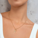 A close-up of a woman wearing a delicate gold chain necklace with two small gold pendants, paired with a white ribbed tank top against a textured gray wall.