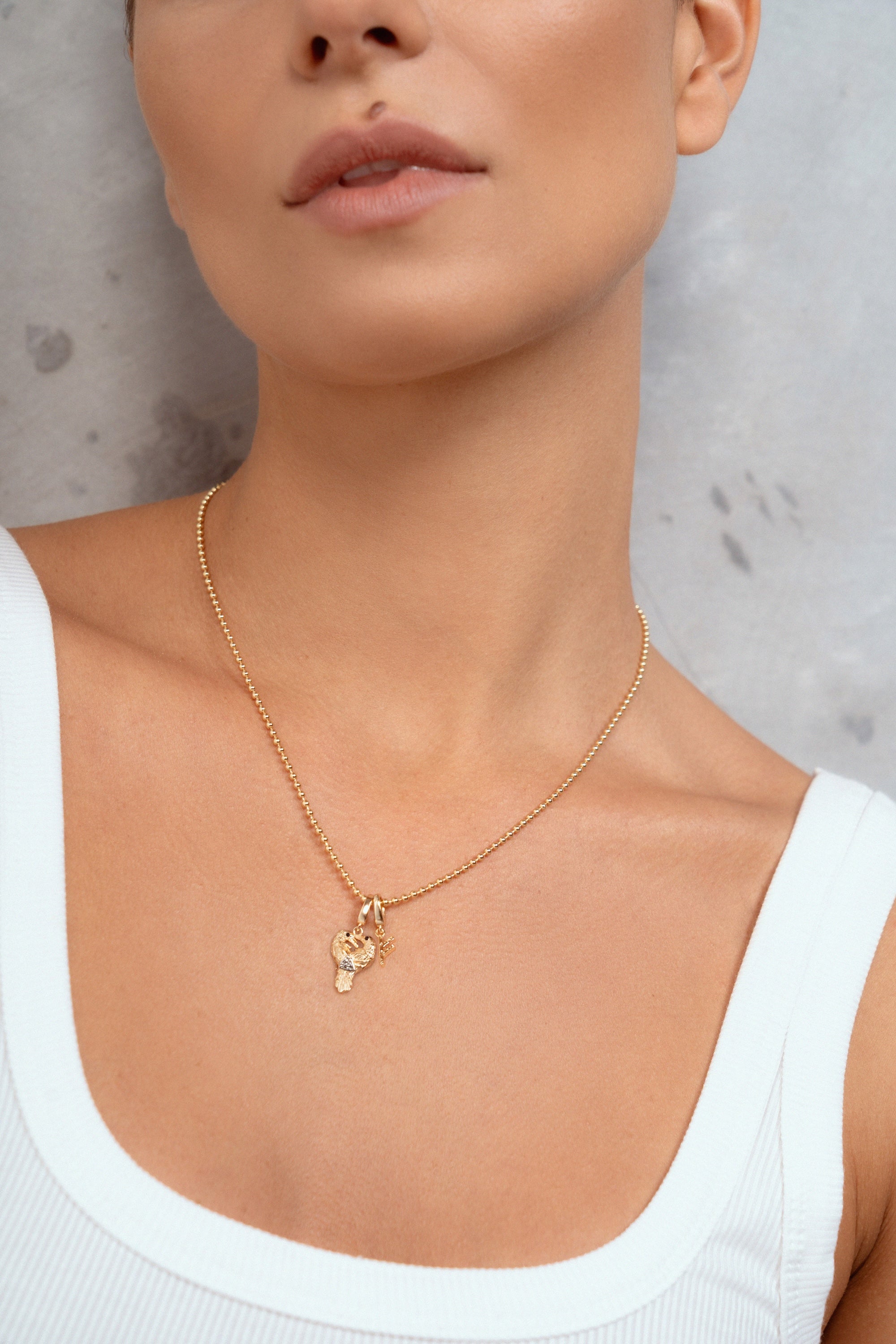 A close-up of a woman wearing a delicate gold chain necklace with two small gold pendants, paired with a white ribbed tank top against a textured gray wall.