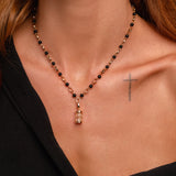 A woman wears a black beaded necklace with an hourglass pendant, a small cross tattoo on her chest, and a black off-the-shoulder top.