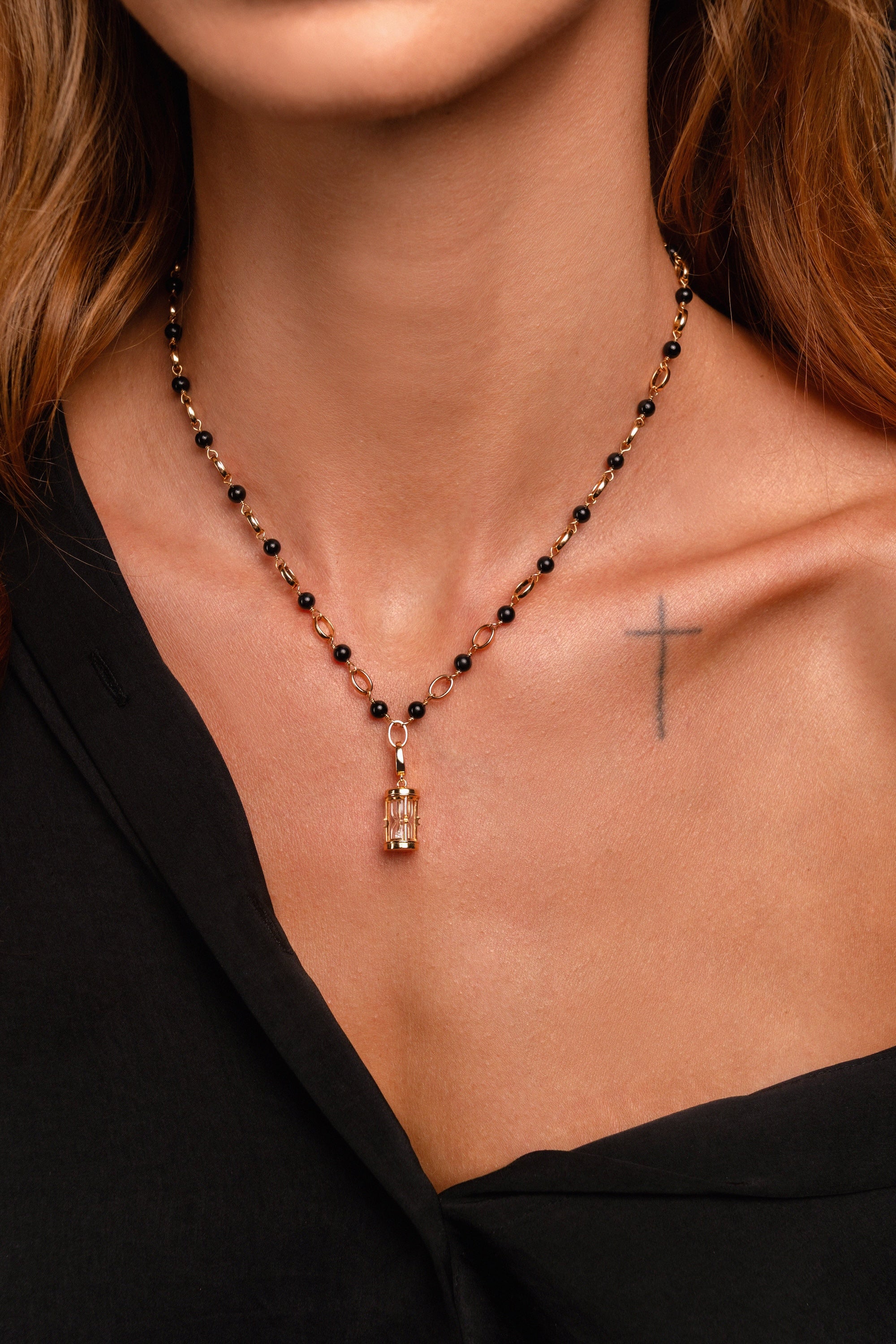 A woman wears a black beaded necklace with an hourglass pendant, a small cross tattoo on her chest, and a black off-the-shoulder top.