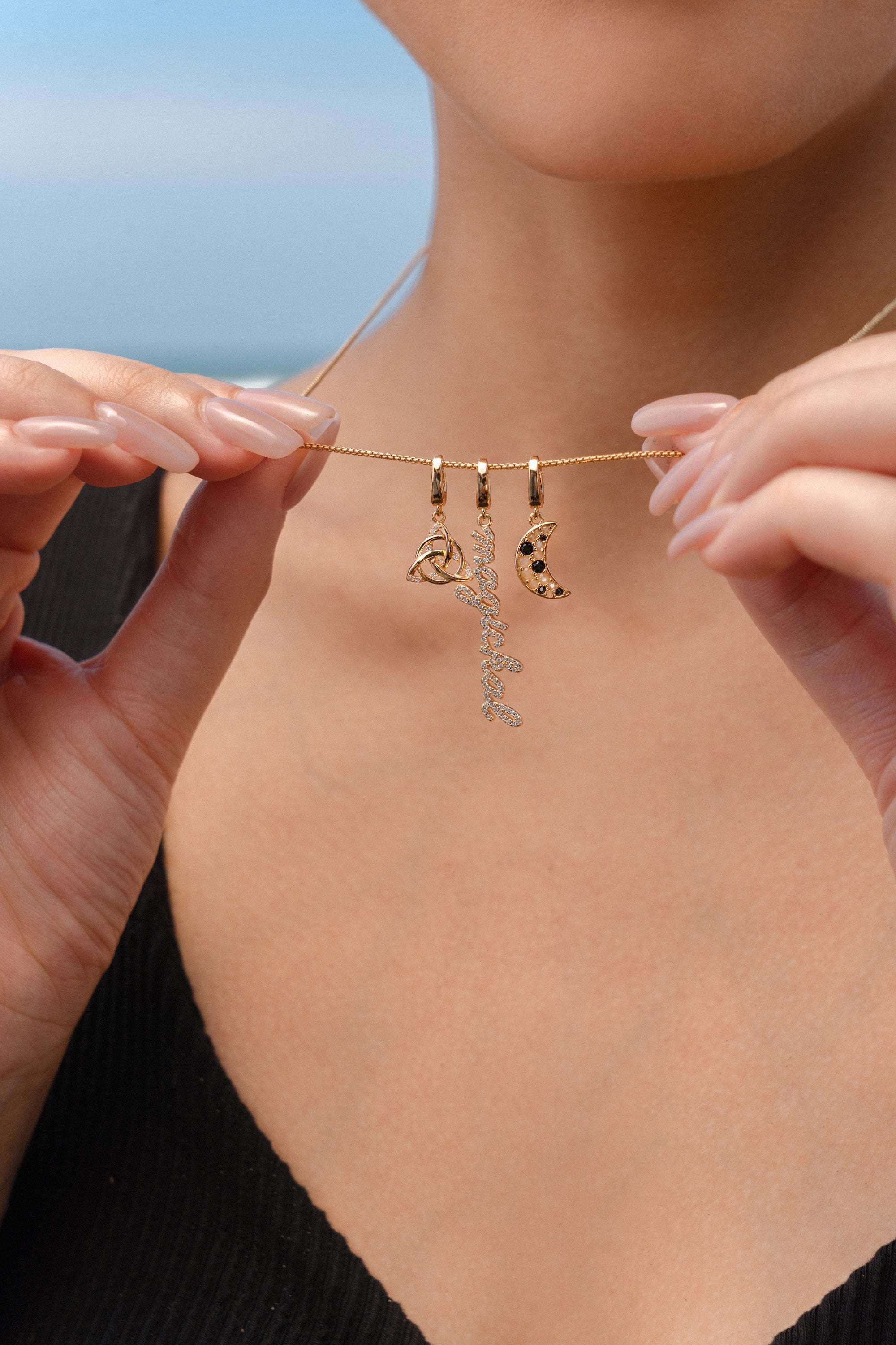 Close-up of a person holding a gold necklace with three charms: a triquetra symbol, a script ‘magickal’ charm, and a gold crescent moon with black and white stones.