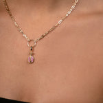 A close-up of a model's neck and chest wearing a delicate gold chain necklace with a small pomegrenate-shaped pendant featuring pink gemstone inlay.