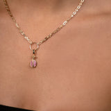A close-up of a model's neck and chest wearing a delicate gold chain necklace with a small pomegrenate-shaped pendant featuring pink gemstone inlay.