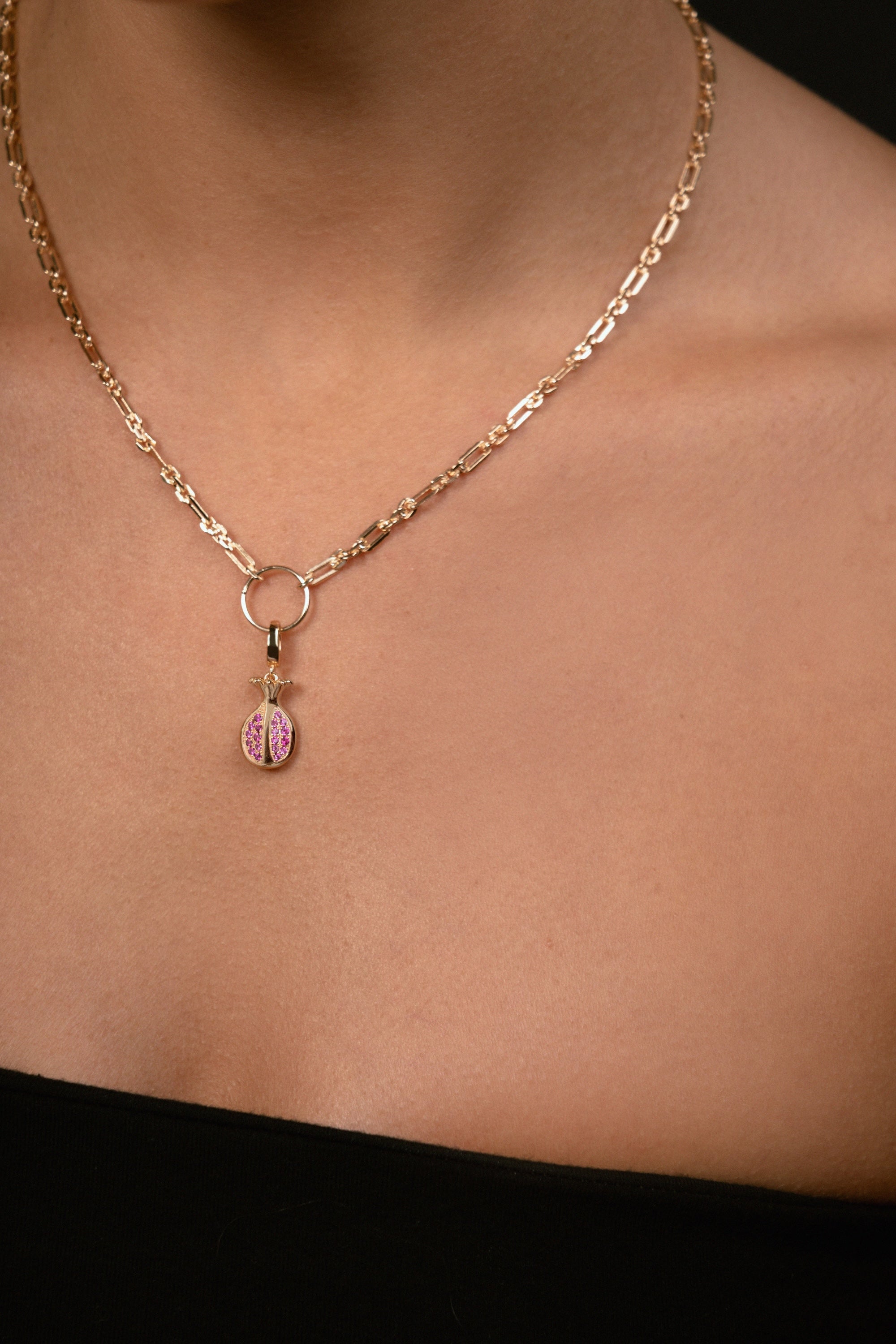 A close-up of a model's neck and chest wearing a delicate gold chain necklace with a small pomegrenate-shaped pendant featuring pink gemstone inlay.